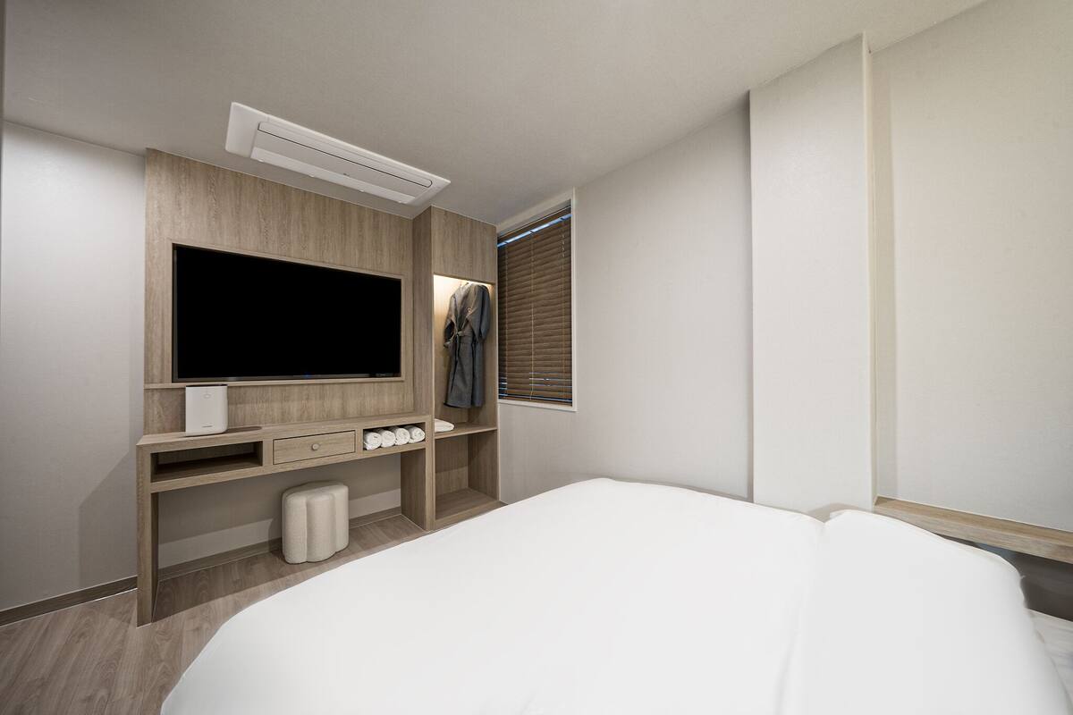 Een moderne hotelkamer met een groot flatscreen televisietoestel, een houten kaptafel, een raam met zonwering, een nachtkastje en een bed.