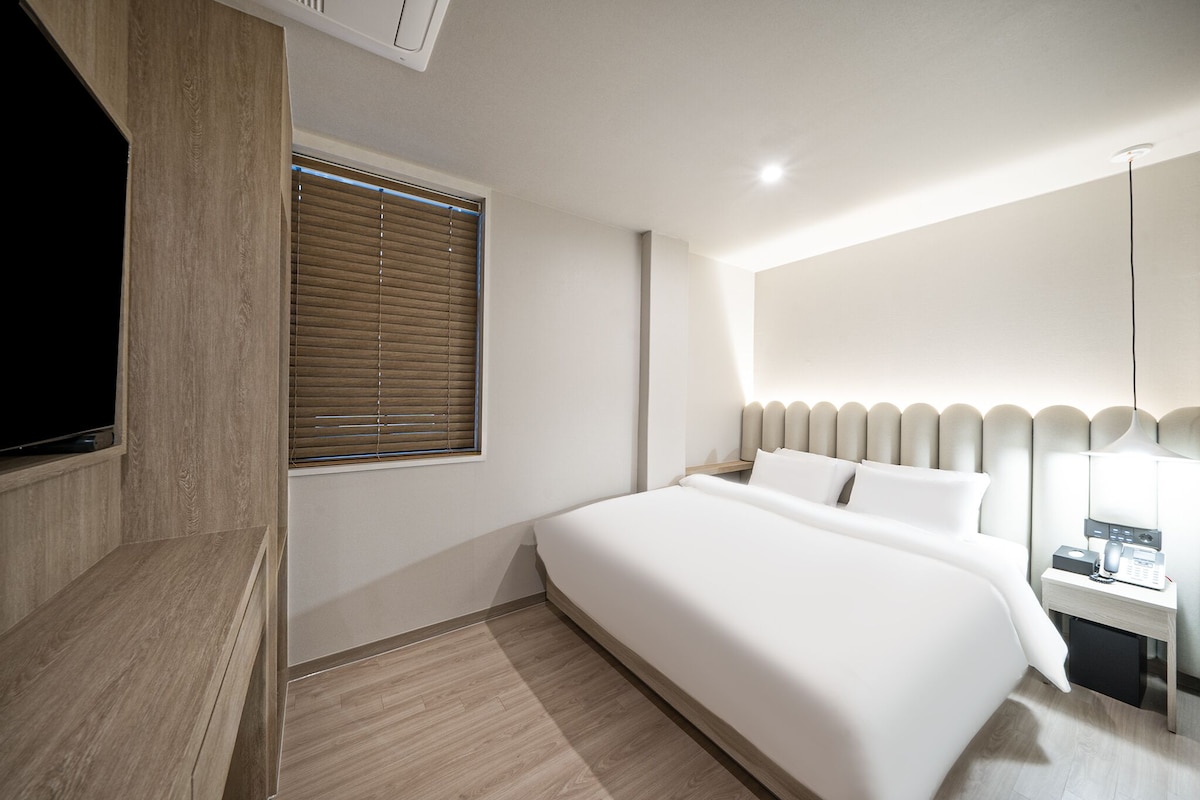A modern hotel room with a large bed, a TV mounted on a wooden panel, a bedside table with a lamp, and a window with blinds.