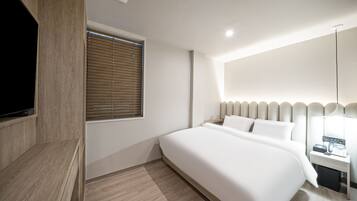 Quarto de hotel moderno com uma cama grande, uma TV fixada em um painel de madeira, uma mesa de cabeceira com abajur e uma janela com persianas.