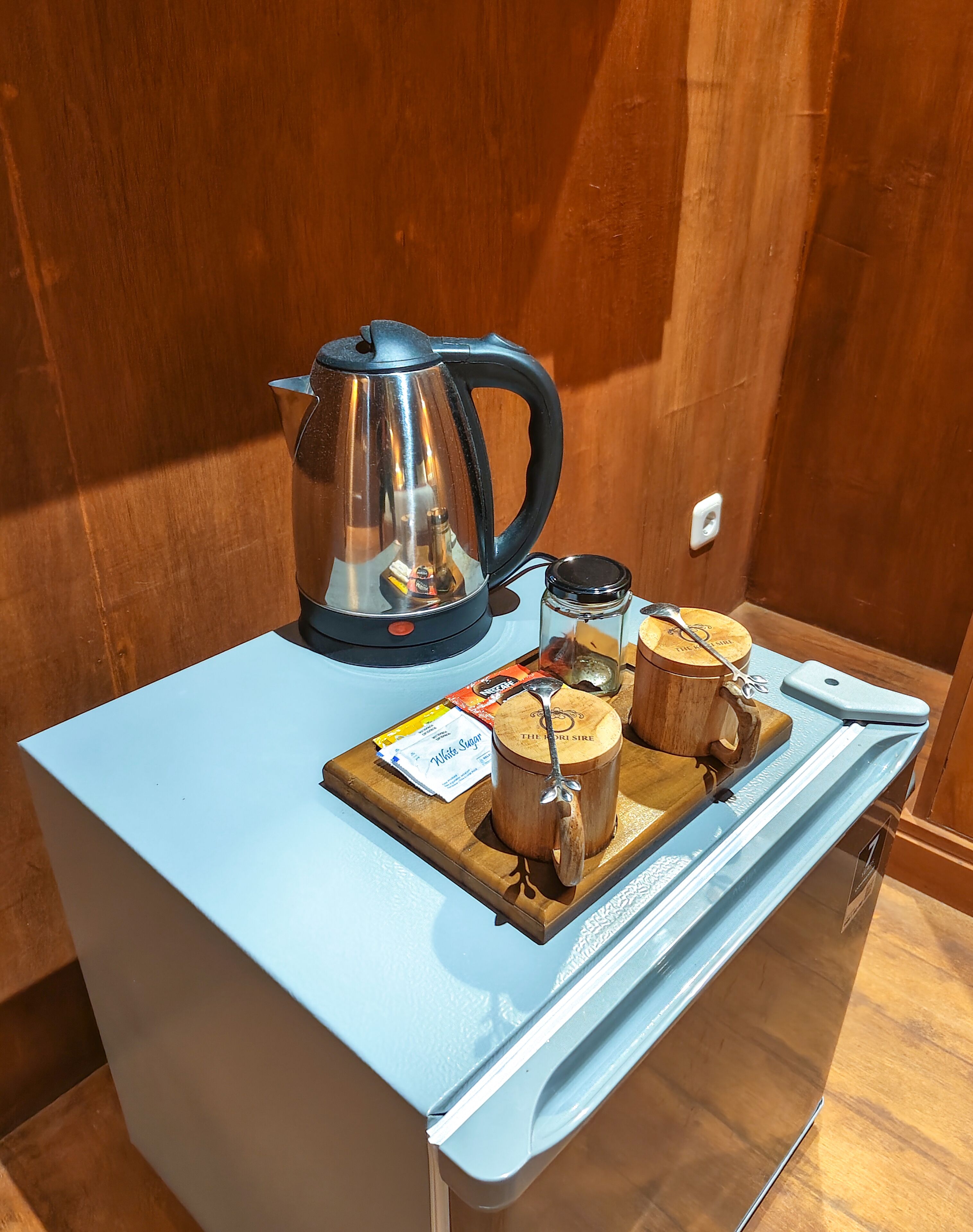 Una kitchenette con un hervidor, una bandeja con tazas de madera y un sobre de azúcar blanca.