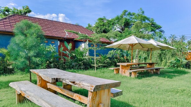 A wooden picnic table with benches in a grassy area, a wooden gazebo with an umbrella, and a house with a red roof in the background. - The Kori Sire (Tanjung)