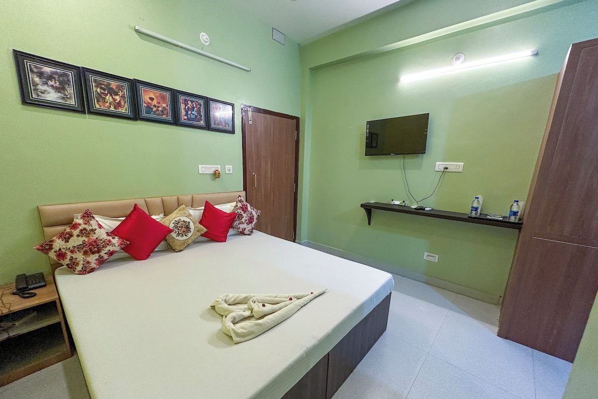Quarto com cama, almofadas, televisão, prateleira com garrafas e quadros na parede.