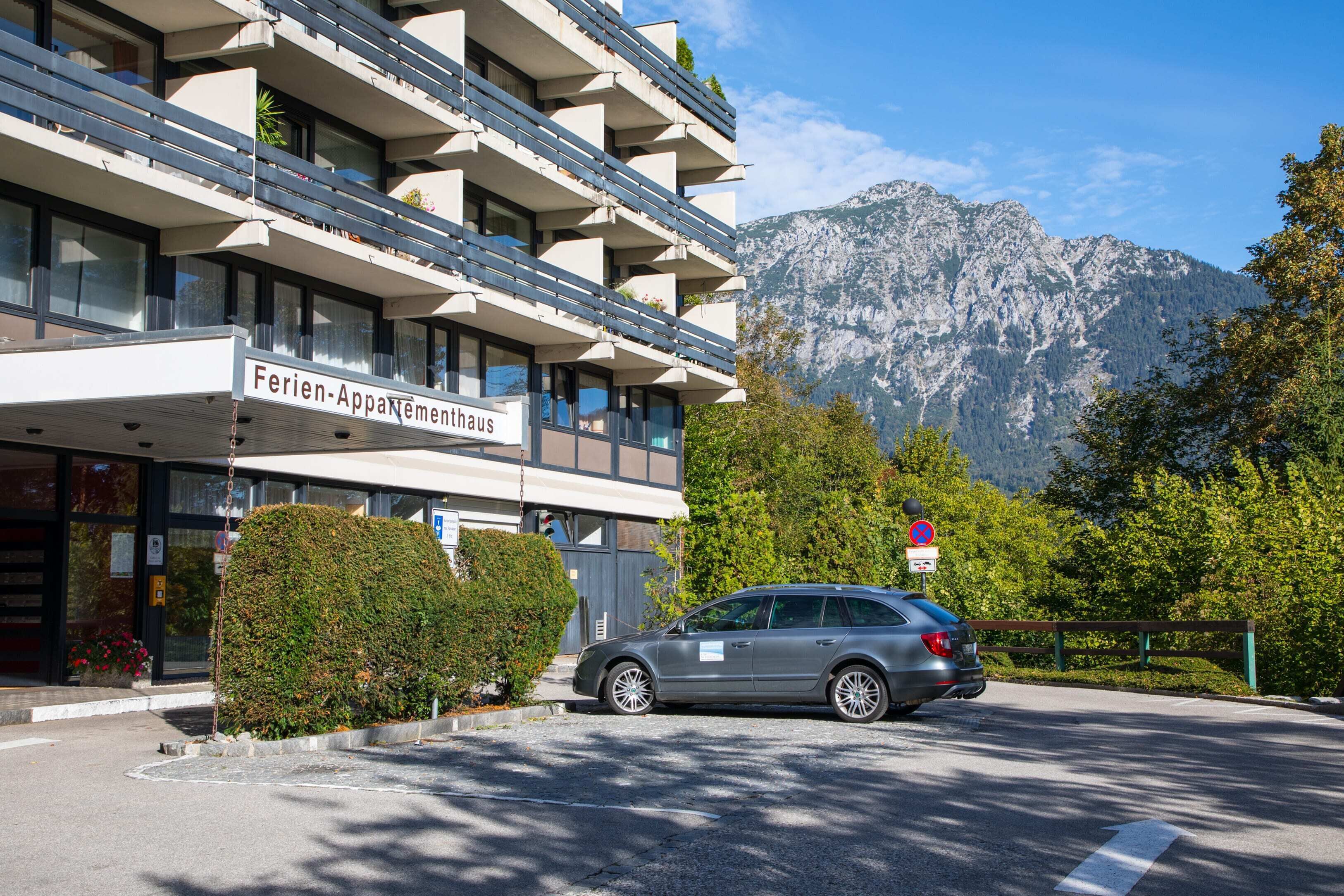 Un hôtel avec une enseigne indiquant « Ferien-Appartementhaus », une voiture garée devant et une chaîne de montagnes en arrière-plan.