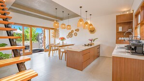 A modern kitchen with a central island, wooden cabinetry, pendant lighting, and a view of the outdoors. - Octo 5 Villa by Betterplace (Canggu)