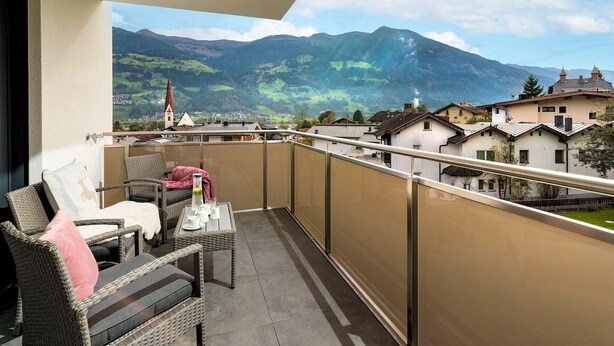 Un balcone con mobili in vimini, un tavolo con tazze e un bicchiere, e vista sulle montagne e sugli edifici.