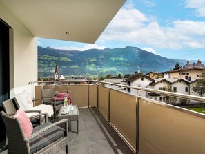 Ein Balkon mit Korbmöbeln, einem Tisch mit Tassen und einem Glas sowie Blick auf Berge und Gebäude.