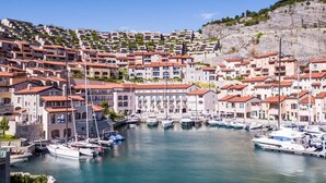 A coastal town with terraced buildings, a marina with boats, and a clear blue sky.