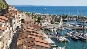 Un pueblo costero con edificios de tejados terracota, un puerto deportivo con numerosos barcos y una vista despejada al mar.