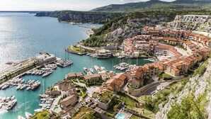 Un pueblo costero con un puerto deportivo, edificios escalonados y montañas al fondo.