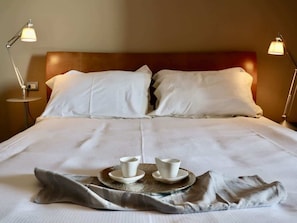 Un letto rifatto con lenzuola bianche, due lampade sui comodini e un vassoio con due tazze e piattini.