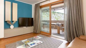 A modern living room with a television, a glass coffee table with magazines, a sofa, and a sliding glass door leading to an outdoor area.