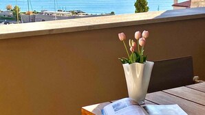 A table with a vase of flowers and an open magazine, overlooking a coastal view with a stone wall and a clear sky.