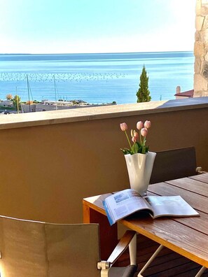 Un tavolo con un vaso di fiori e una rivista aperta, affacciato su una vista costiera con un muro di pietra e cielo sereno.