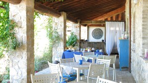 An outdoor patio with stone walls, wooden beams, and white chairs. Tables are covered with blue tablecloths, and there's a large clock on the wall. - Villa Profidia (Gualdo Cattaneo)