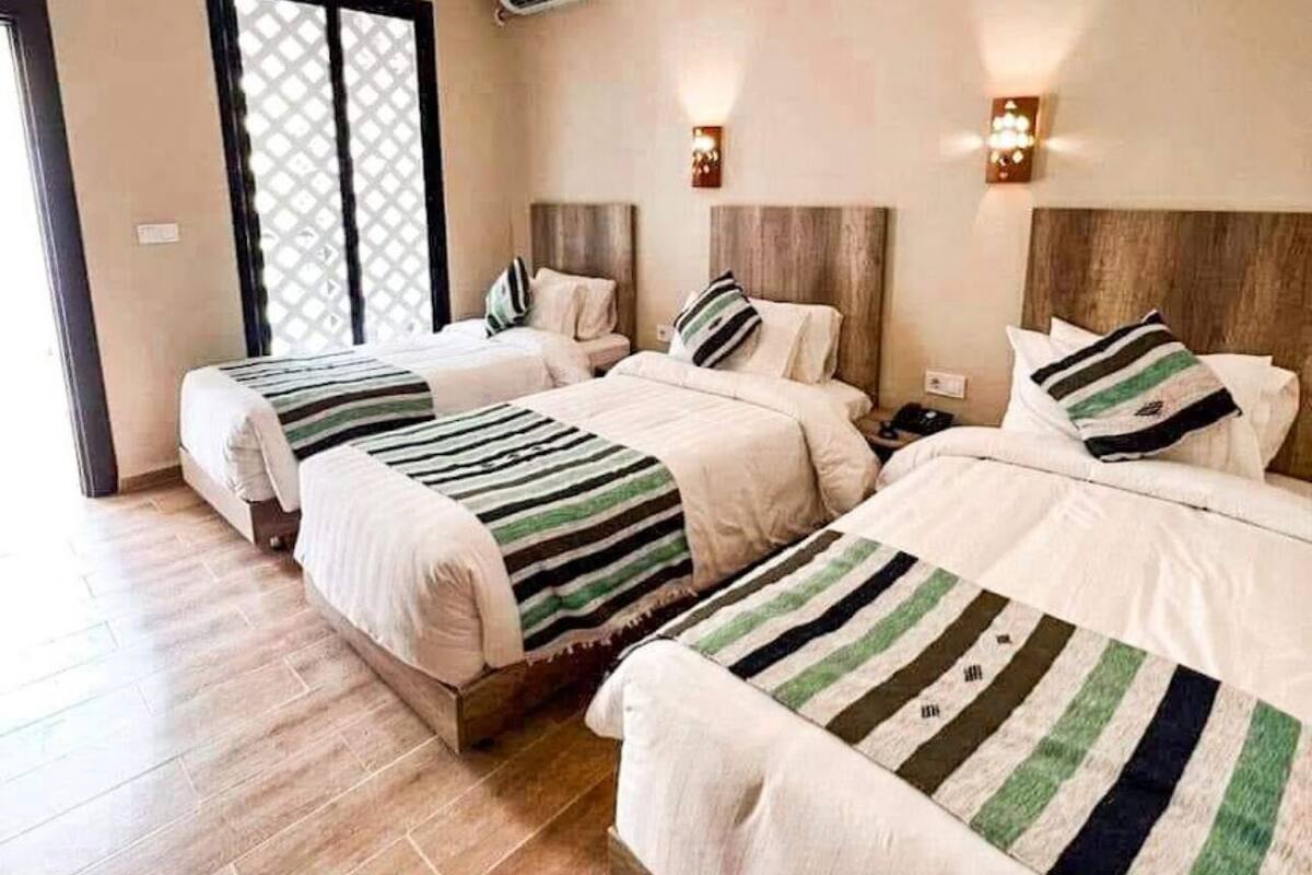 A hotel room with three beds, a wooden floor, a window with a lattice design, and an air conditioning unit on the wall.