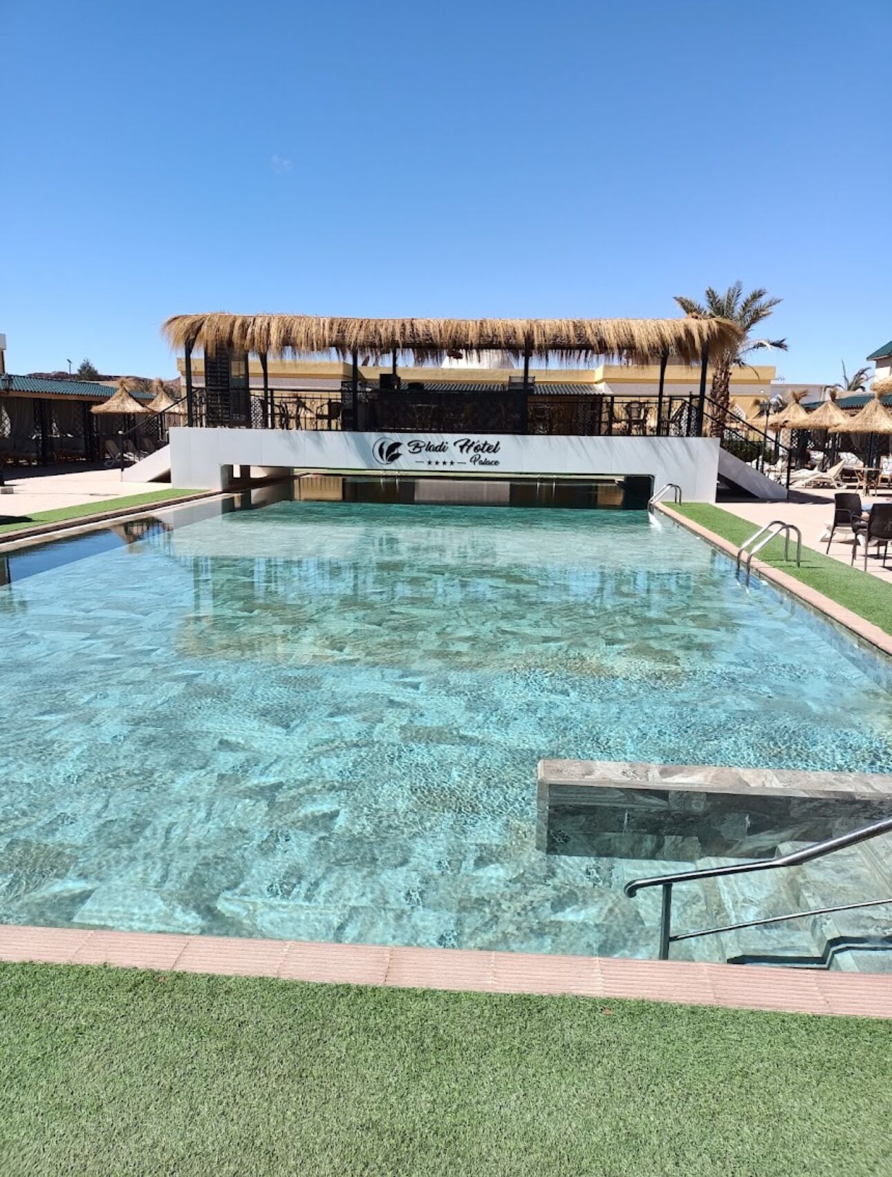 A clear pool with a sign reading "Bladi Hotel Palace" and a thatched roof structure in the background.