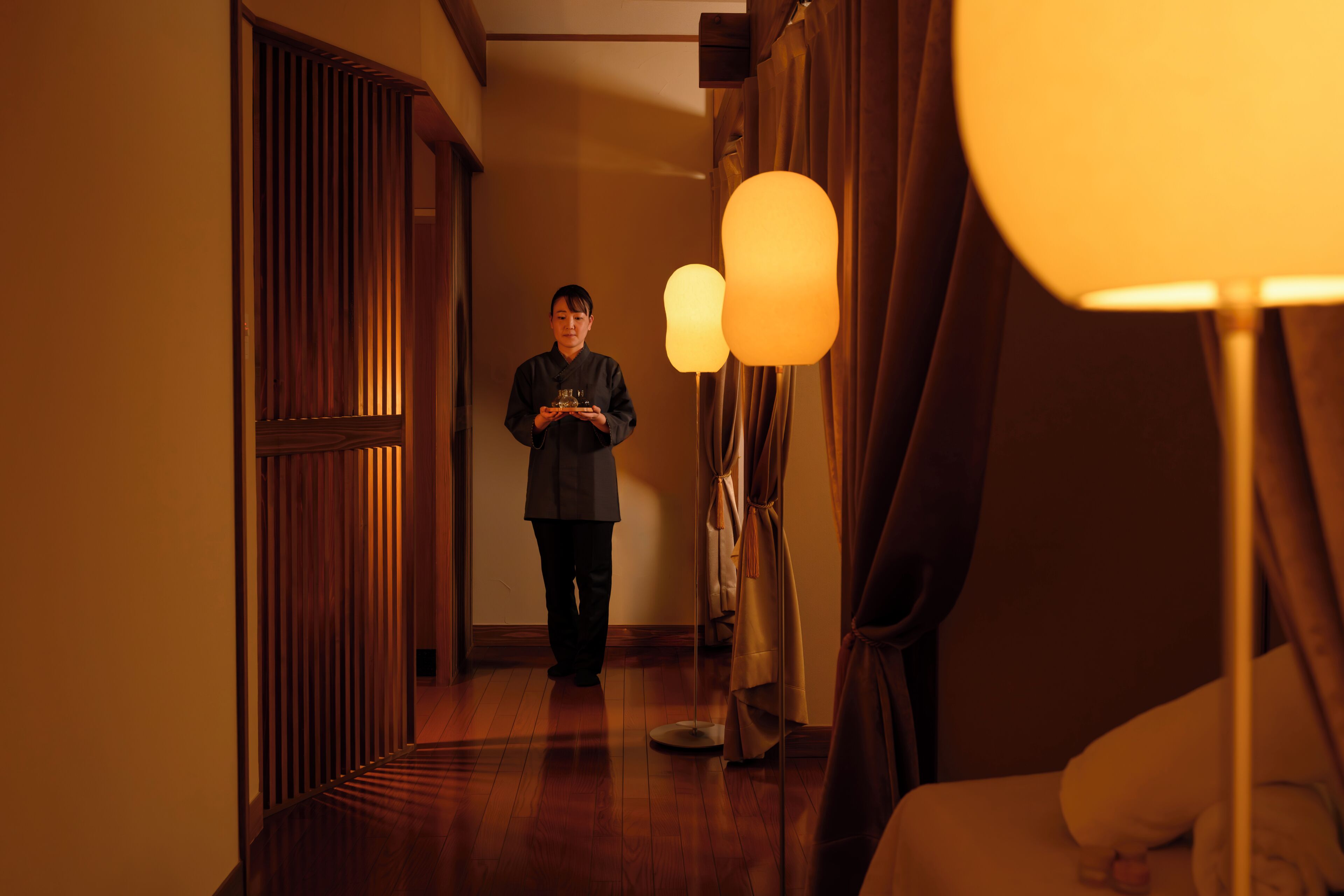 A hotel corridor with wooden paneling, a person holding a tray, and a bed with a headboard.