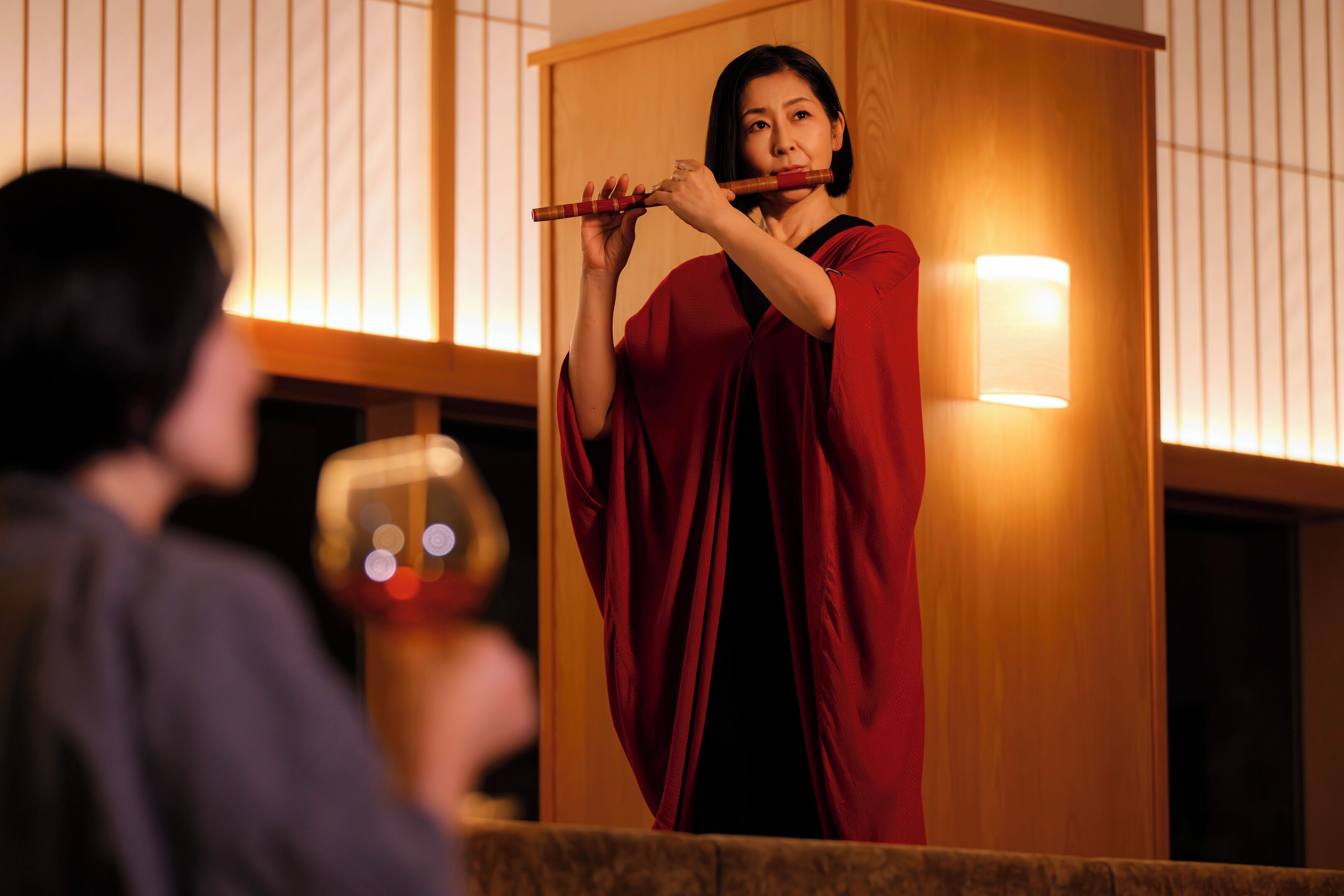 A person playing a flute inside a room with wooden paneling and a lit wall lamp.