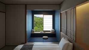 Camera d'albergo con un letto grande, una finestra vista verde, un piccolo angolo relax con un cuscino, e uno scaffale a parete con libri e un vaso.