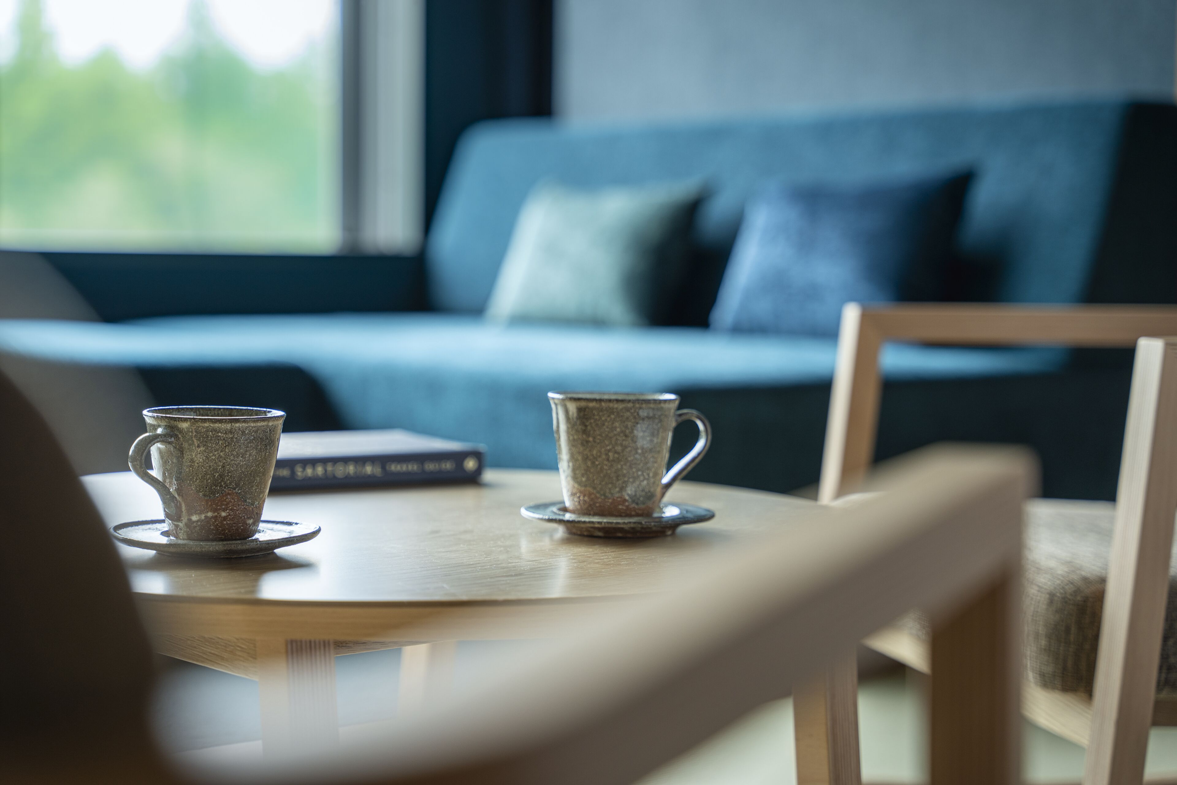 A table with two ceramic cups and a book, a wooden chair, and a blue sofa in the background.