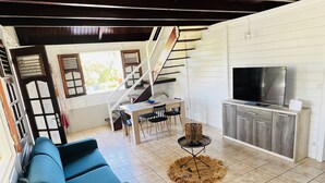 A spacious living area with a blue sofa, a dining table with chairs, a TV on a wooden cabinet, and a staircase leading to an upper level.