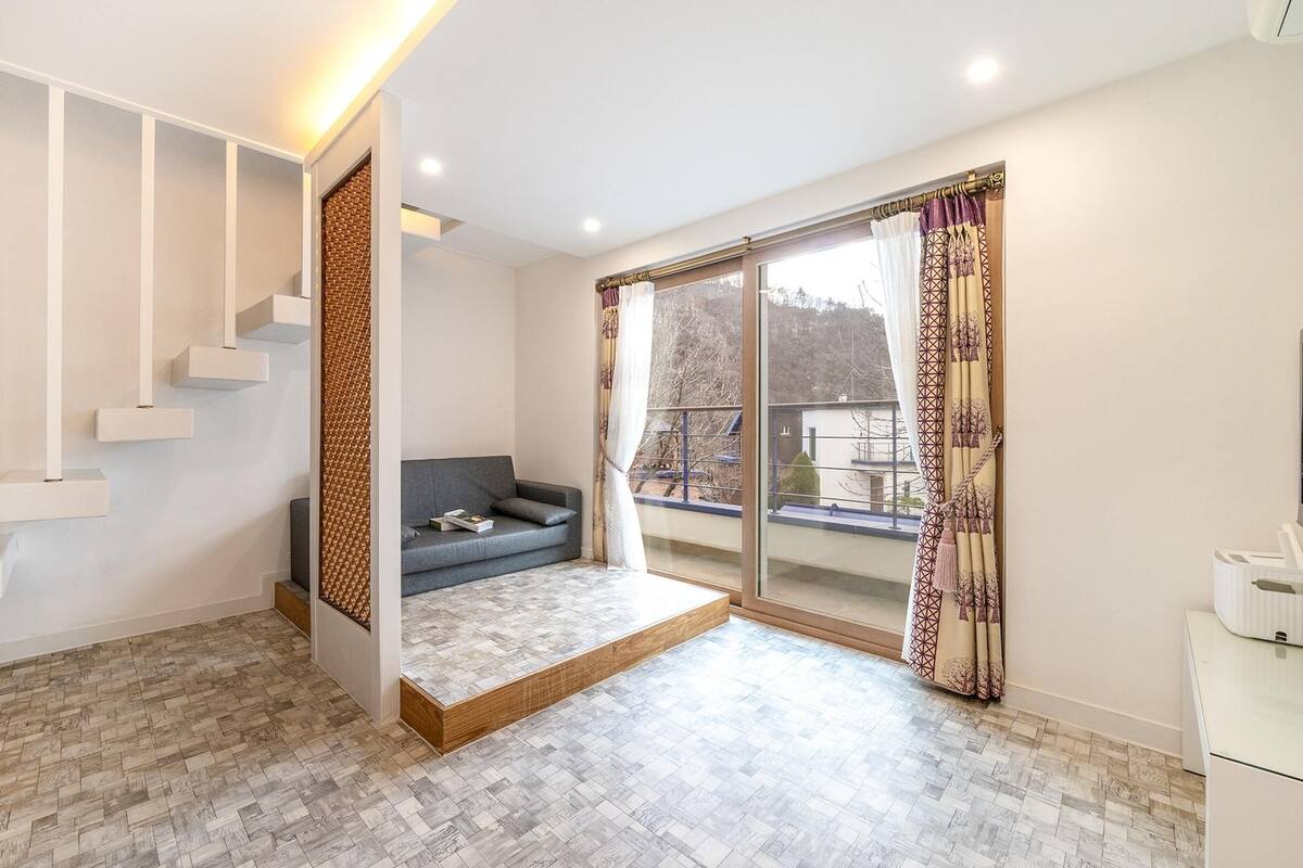 Ein modernes Wohnzimmer mit einer eingebauten Treppe, einem grauen Sofa und einem großen Fenster mit gemusterten Vorhängen.