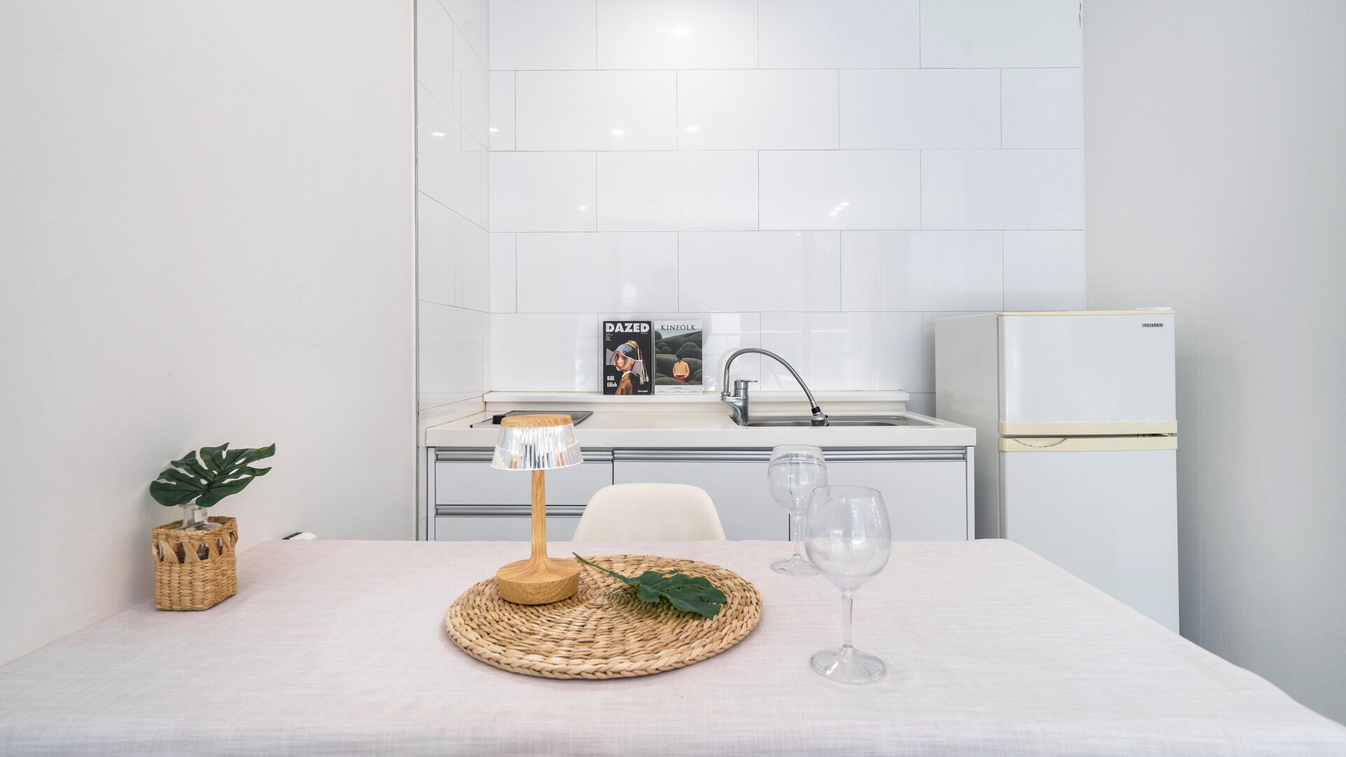 A modern kitchen with white tiles, a white refrigerator, a sink, and a dining table with a placemat, a glass, and a plant.