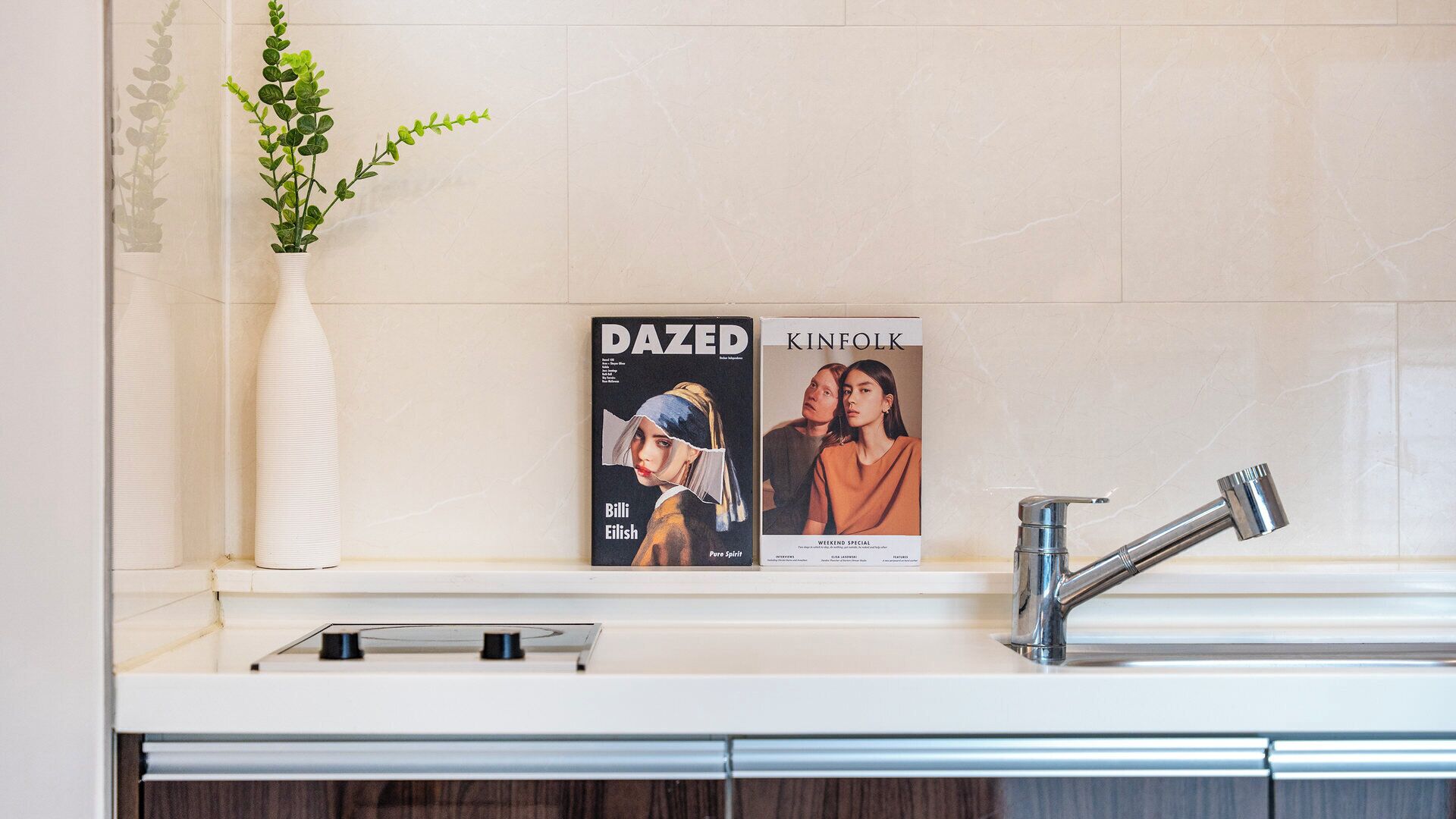 A bathroom with a white sink, a modern faucet, and a magazine titled 'DAZED' and 'KINFOLK' on the wall.