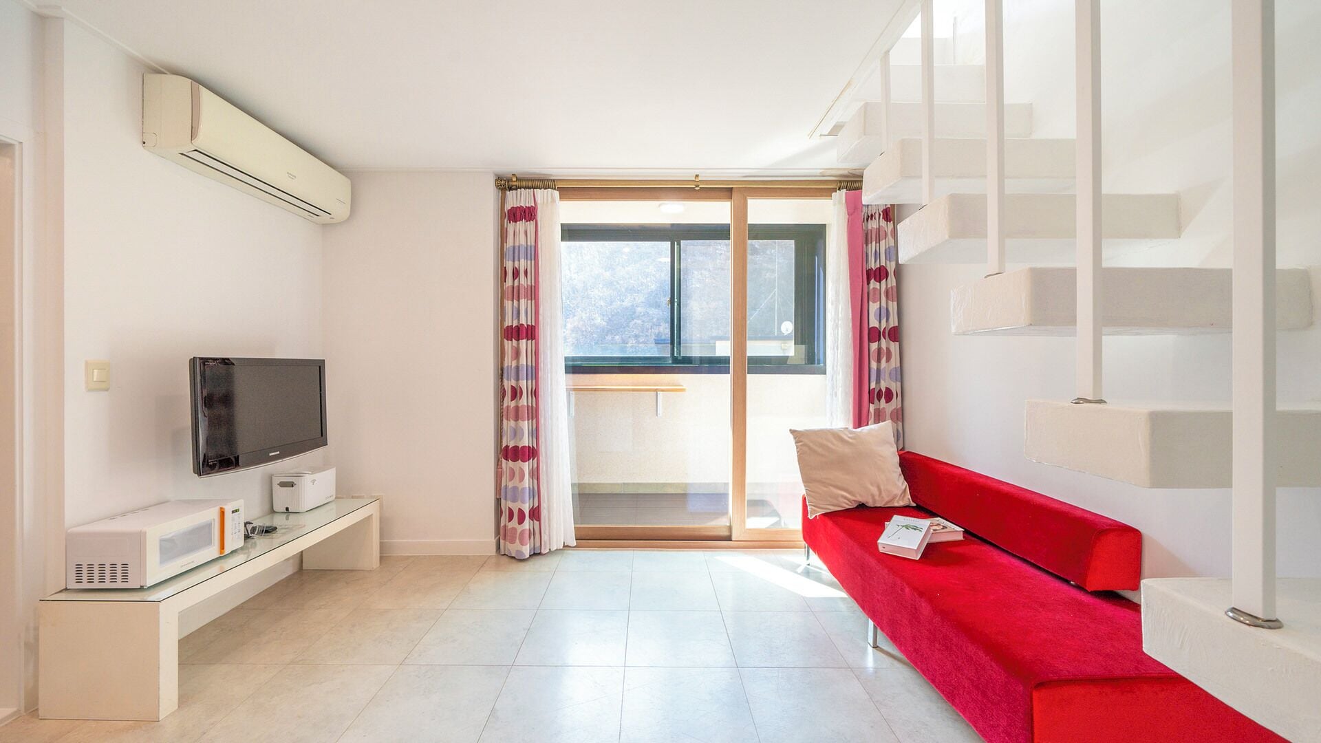 A modern living room with a flat-screen TV, a small white cabinet, a red sofa, and a sliding glass door leading to a balcony.