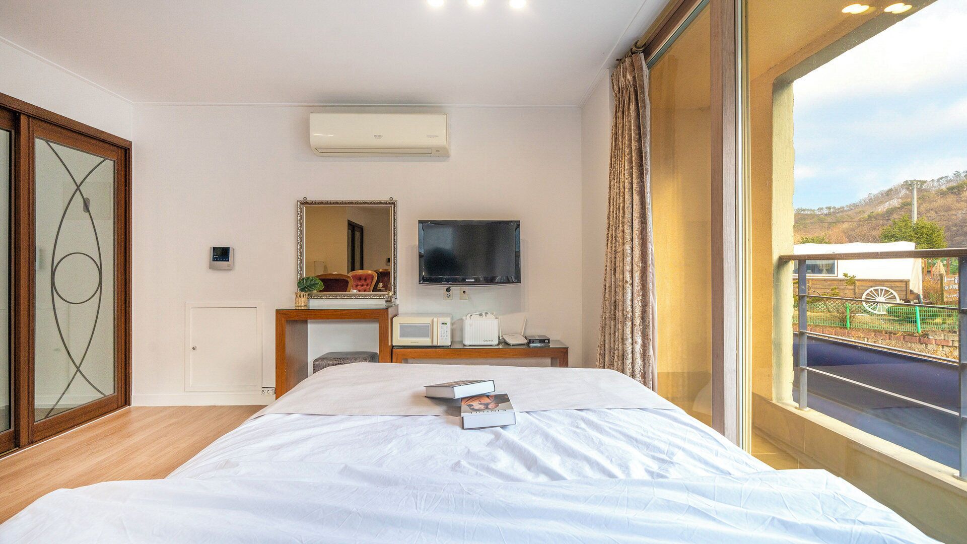 A hotel room with a bed, a television, a desk with a microwave, and a window with a view of the outdoors.