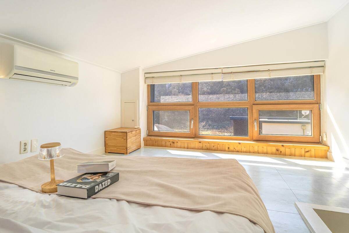 Ein Schlafzimmer mit großem Fenster, einem Bett mit beiger Decke, einem Nachttisch mit Lampe und einem Buch.