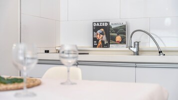 A modern kitchen with a DaZed Kinfolk magazine on the wall, a stainless steel faucet, and a table with glasses.