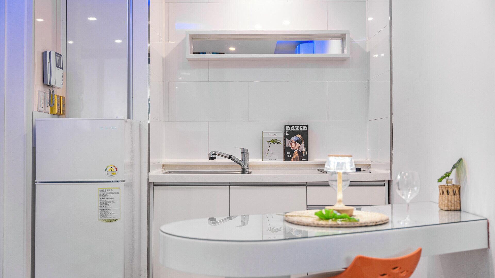 Uma cozinha moderna com geladeira branca, pia e área de refeições com mesa de vidro e uma cadeira.