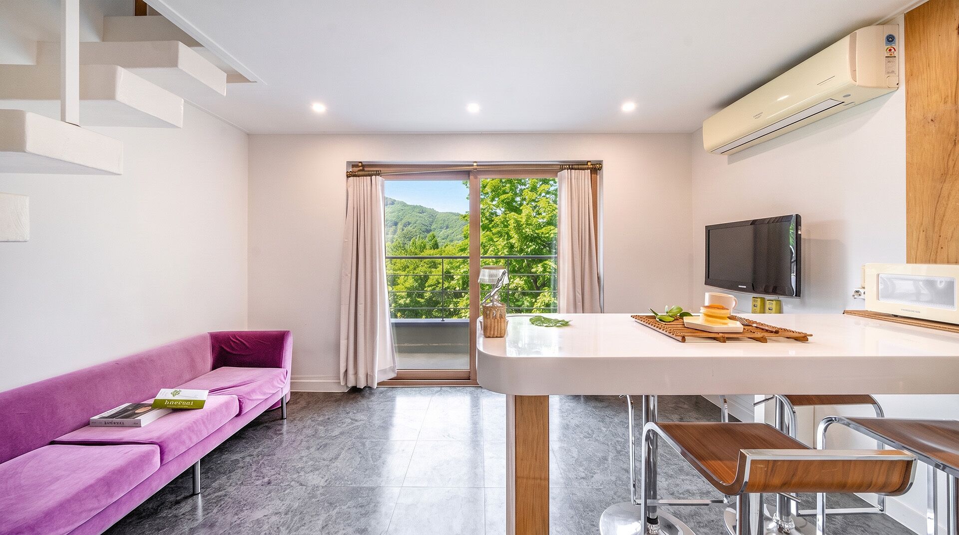 A modern kitchen with a flat-screen TV, a bar area with stools, a purple sofa, and a view of greenery through the window.