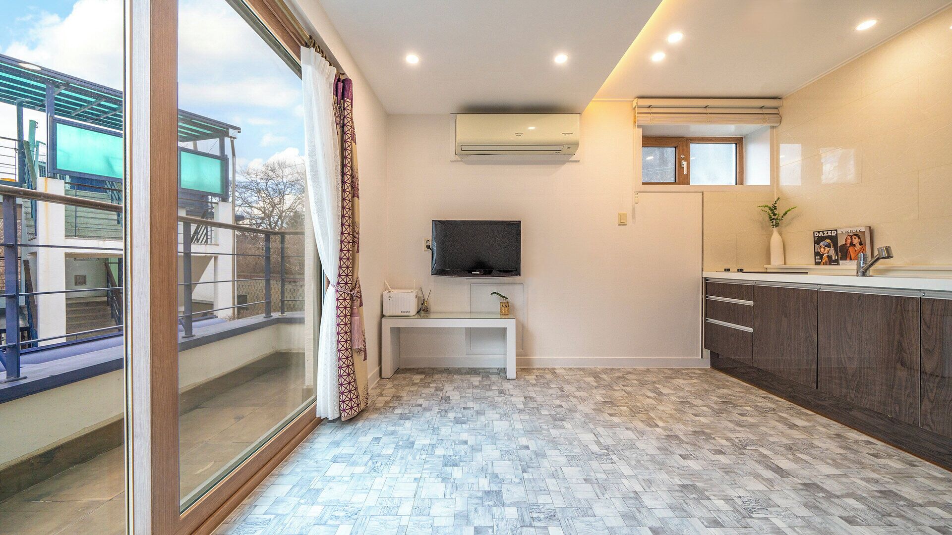 A modern kitchen with a built-in stove, a flat-screen TV, and a window overlooking a balcony.