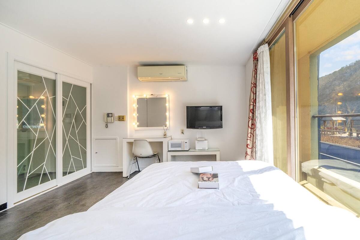 Ein Hotelzimmer mit einem Bett, einem Schreibtisch mit Computer, einem Fernseher und einem großen Fenster mit Blick auf einen Balkon und die Umgebung.