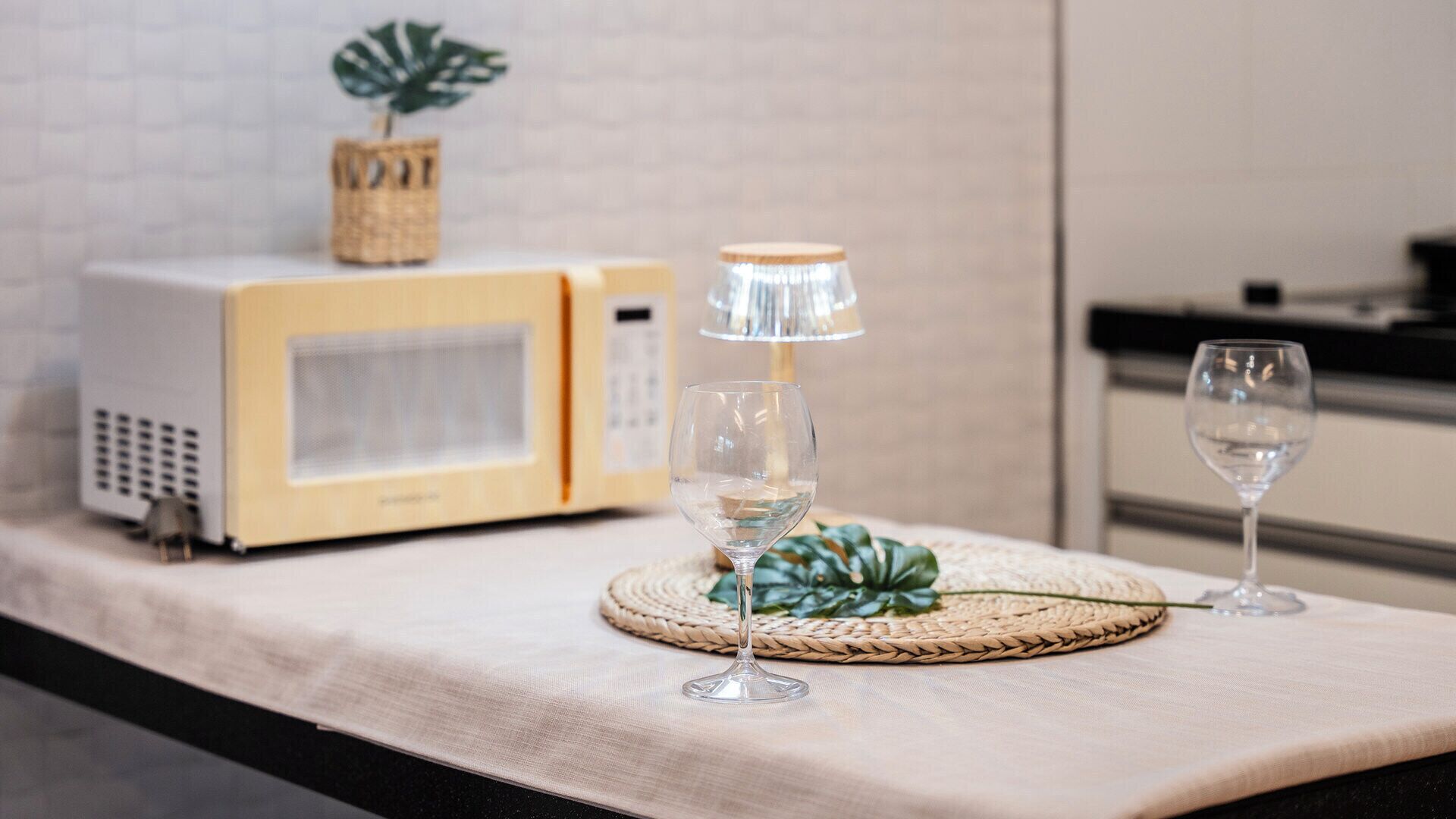 A kitchen counter with a microwave, a glass lamp, and two wine glasses on woven placemats.