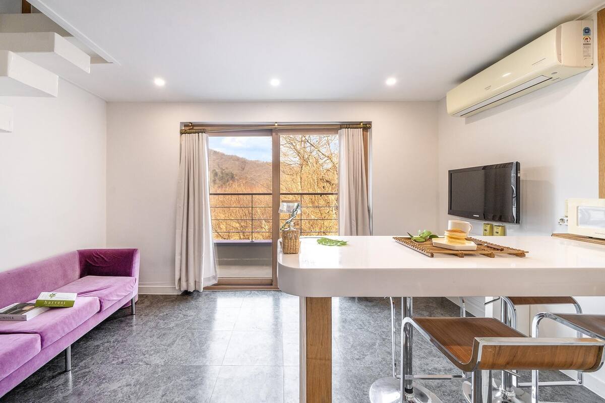 Eine moderne Küche mit Essbereich, einem violetten Sofa und Blick auf Herbstbäume durch das Fenster.