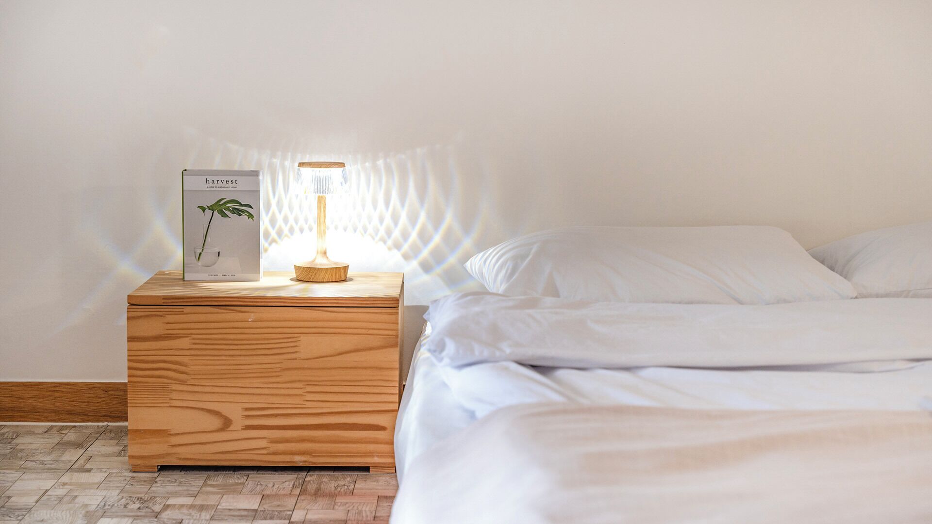 Una mesita de noche de madera con una lámpara y un libro titulado 'harvest' al lado de una cama con ropa de cama blanca.