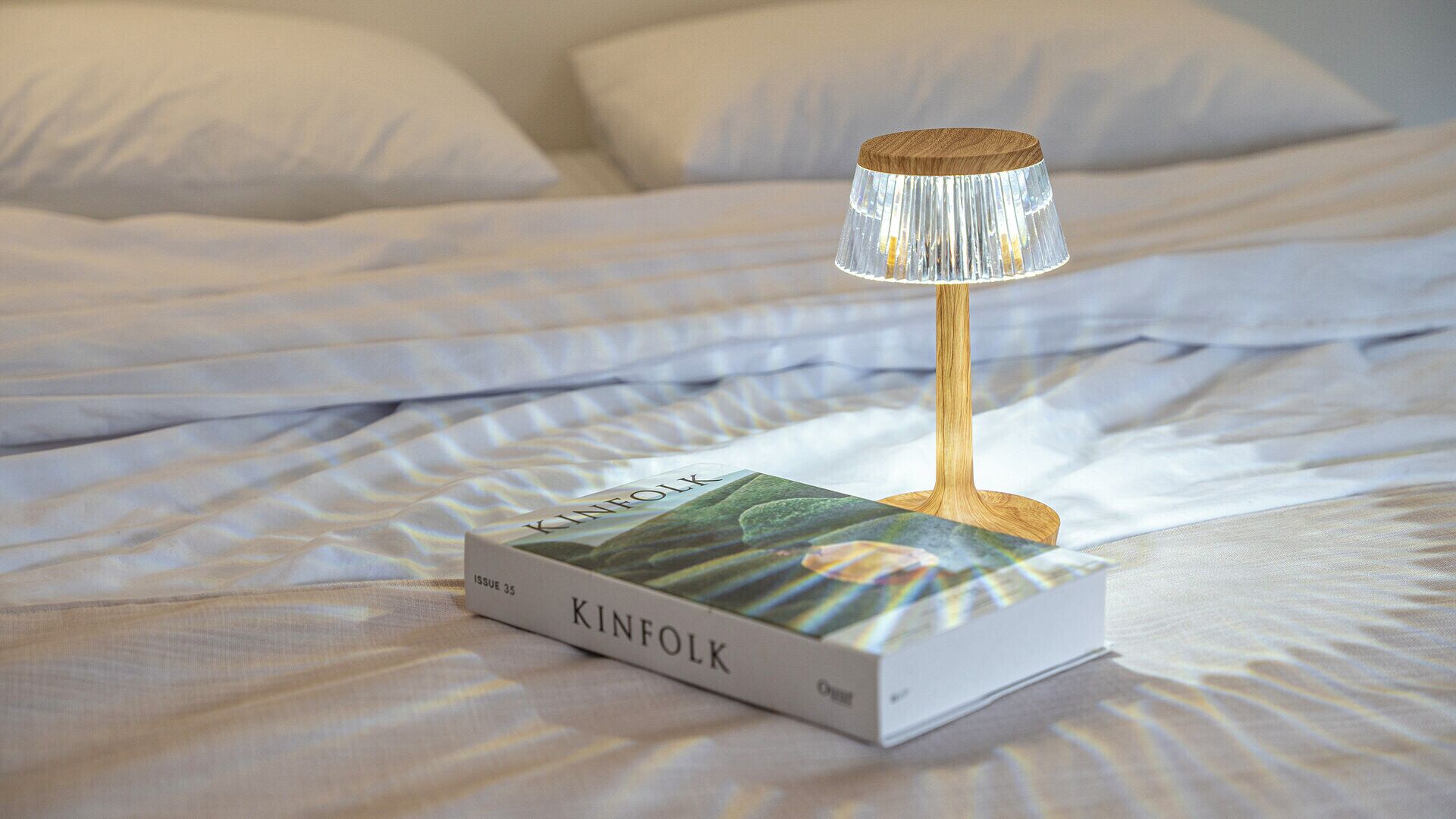 A book titled 'KINFOLK ISSUE 35' on a bed with white bedding and a wooden lamp with a pleated lampshade.