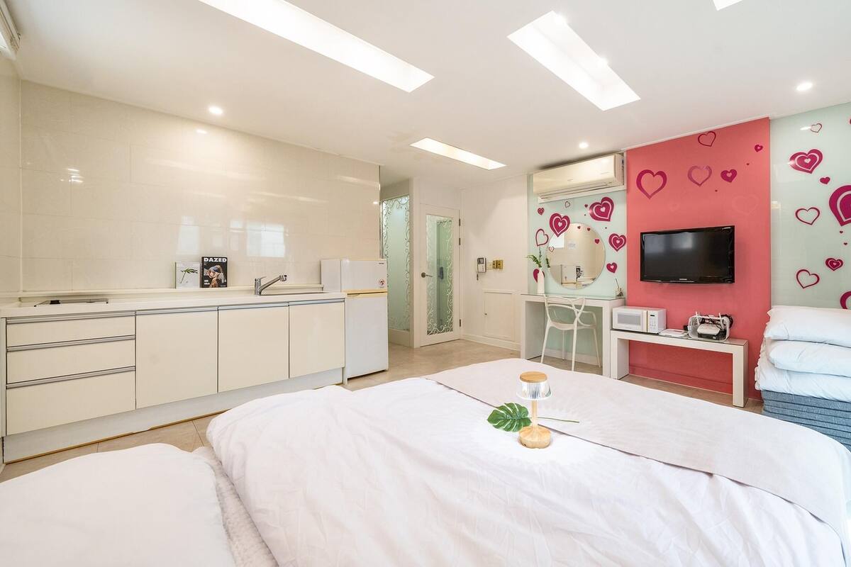 Ein modernes Schlafzimmer mit einem großen Bett, einem Schminktisch mit Spiegel, einem Fernseher und Wanddekorationen mit Herzmotiven.