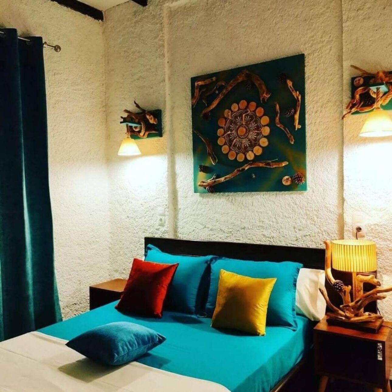 Una cama con almohadas azules y amarillas, una pared con una obra de arte circular y una mesita de noche de madera con una lámpara.
