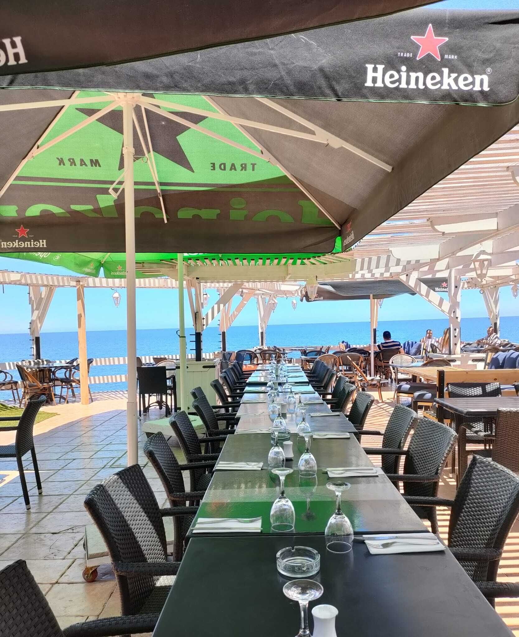 Een buitenterras met tafels en stoelen, een uitzicht op de zee en parasols met het Heineken logo.