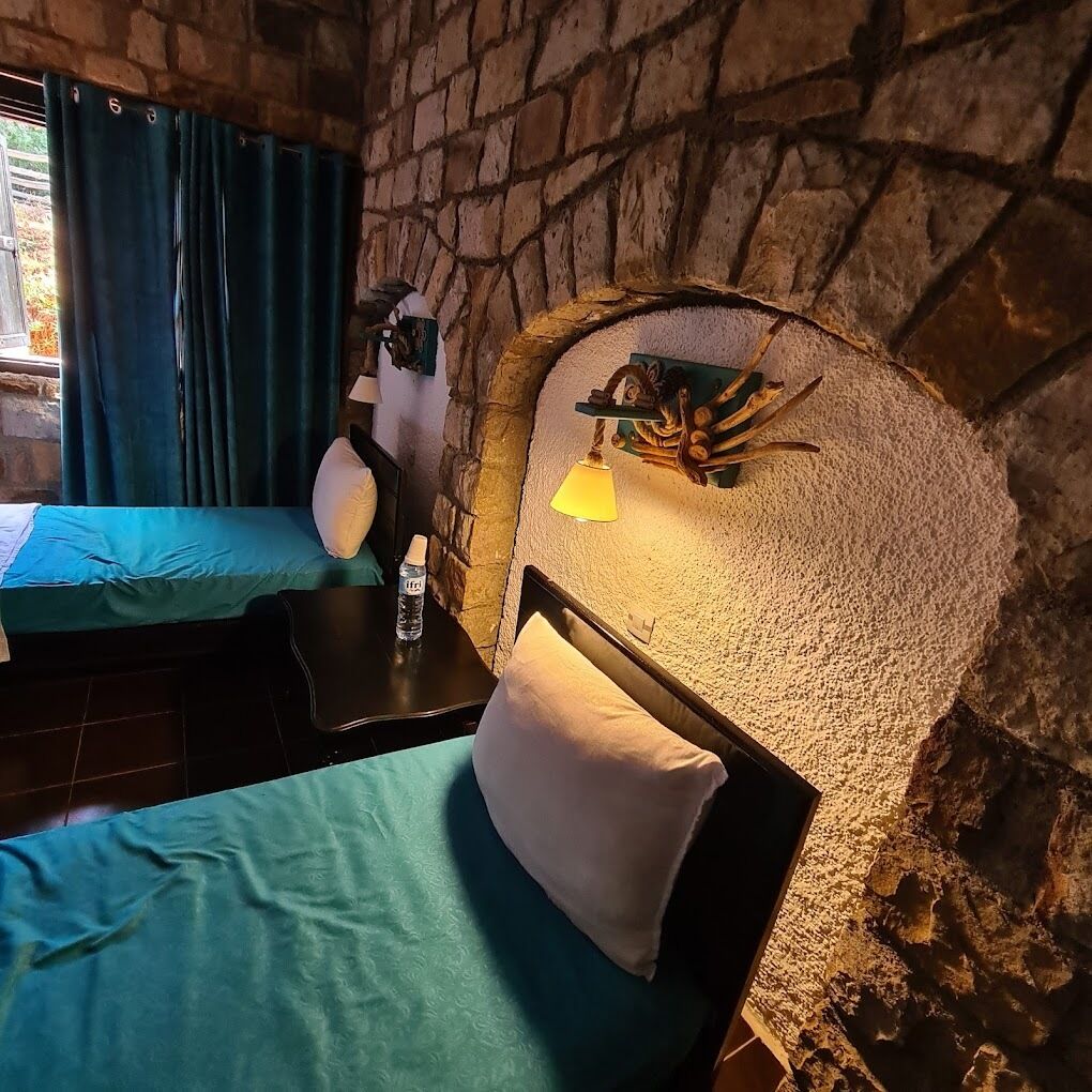 Een kamer met twee bedden, elk met een blauwgroene sprei en witte kussens. Er staat een klein tafeltje met een waterfles en een lamp. De muren zijn van steen, en er is een raam met blauwe gordijnen.