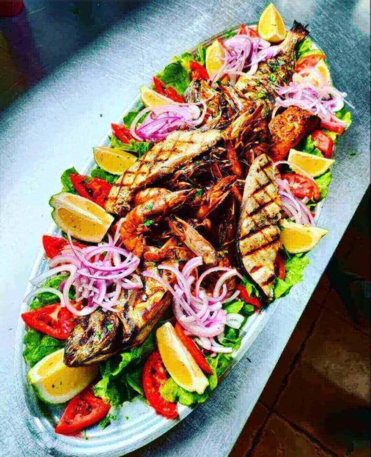 A platter of grilled fish with lemon slices, tomatoes, and red onions.