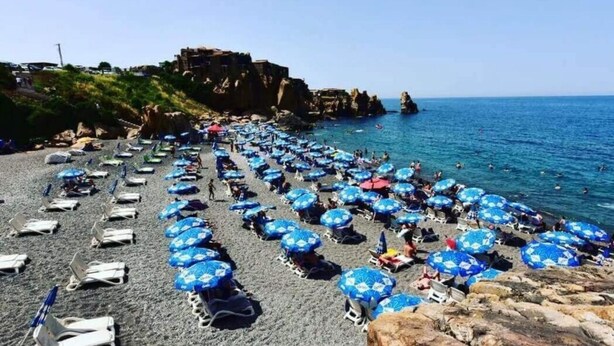A beach with rows of sun loungers and umbrellas, rocky cliffs in the background, and a clear blue sea. - Auberge Thais (Toudja)