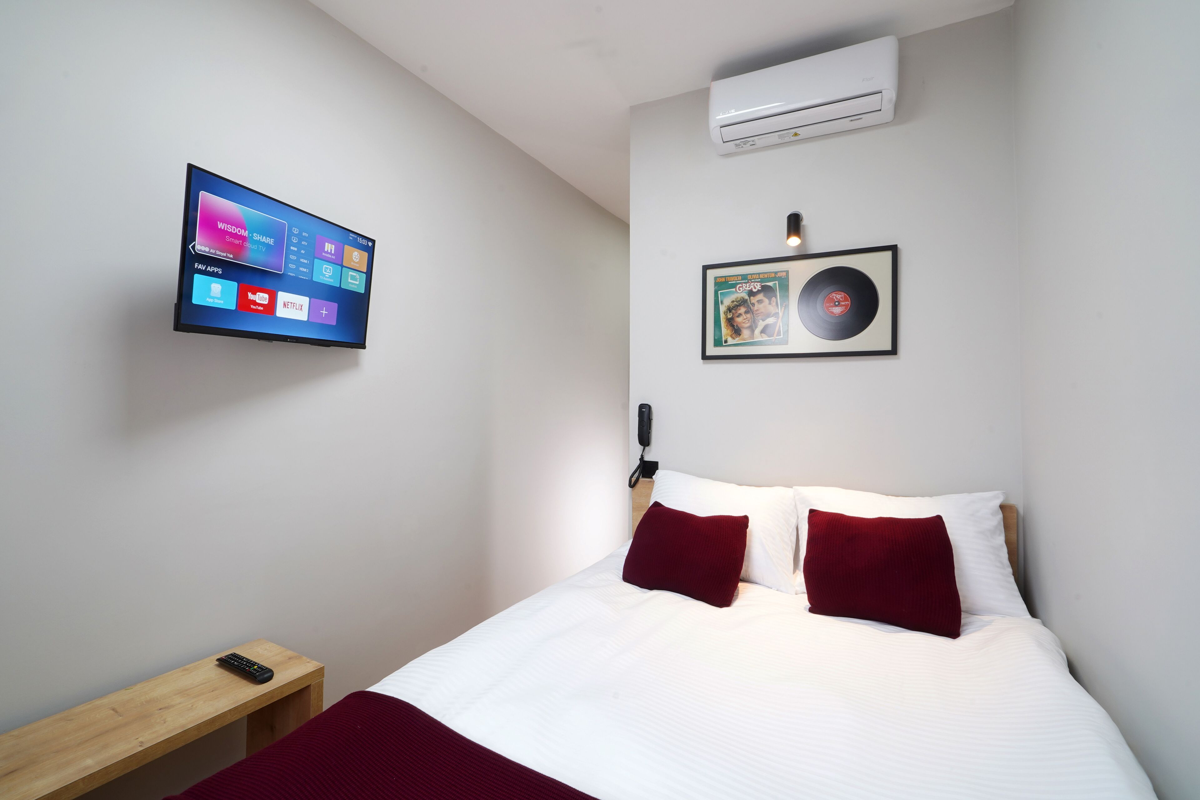 A hotel room with a bed, a bedside table, a TV mounted on the wall, an air conditioner, and a framed picture on the wall.
