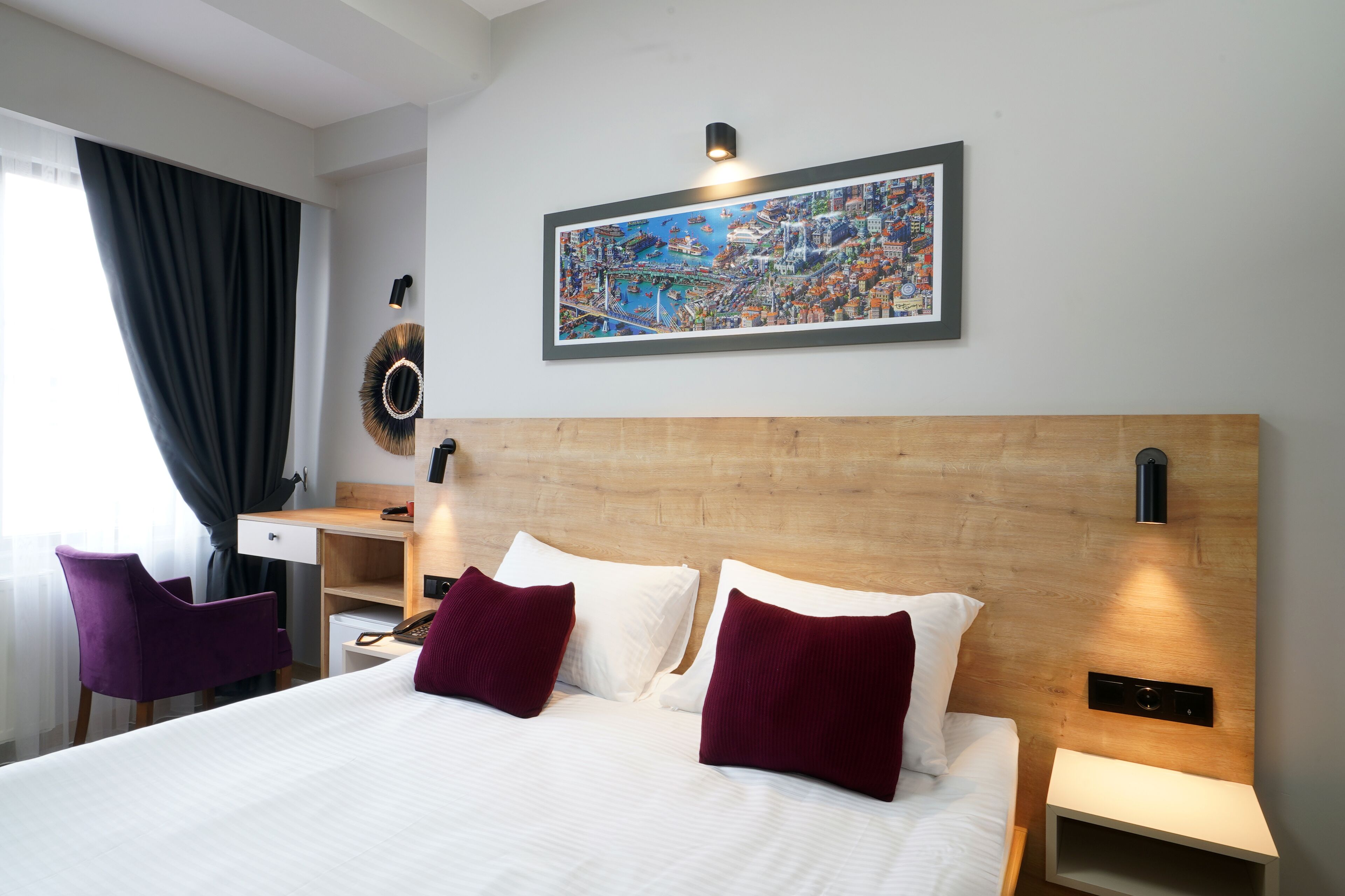 A hotel room with a large bed, two pillows, a desk, a chair, a nightstand, a wall-mounted lamp, and a framed cityscape picture.