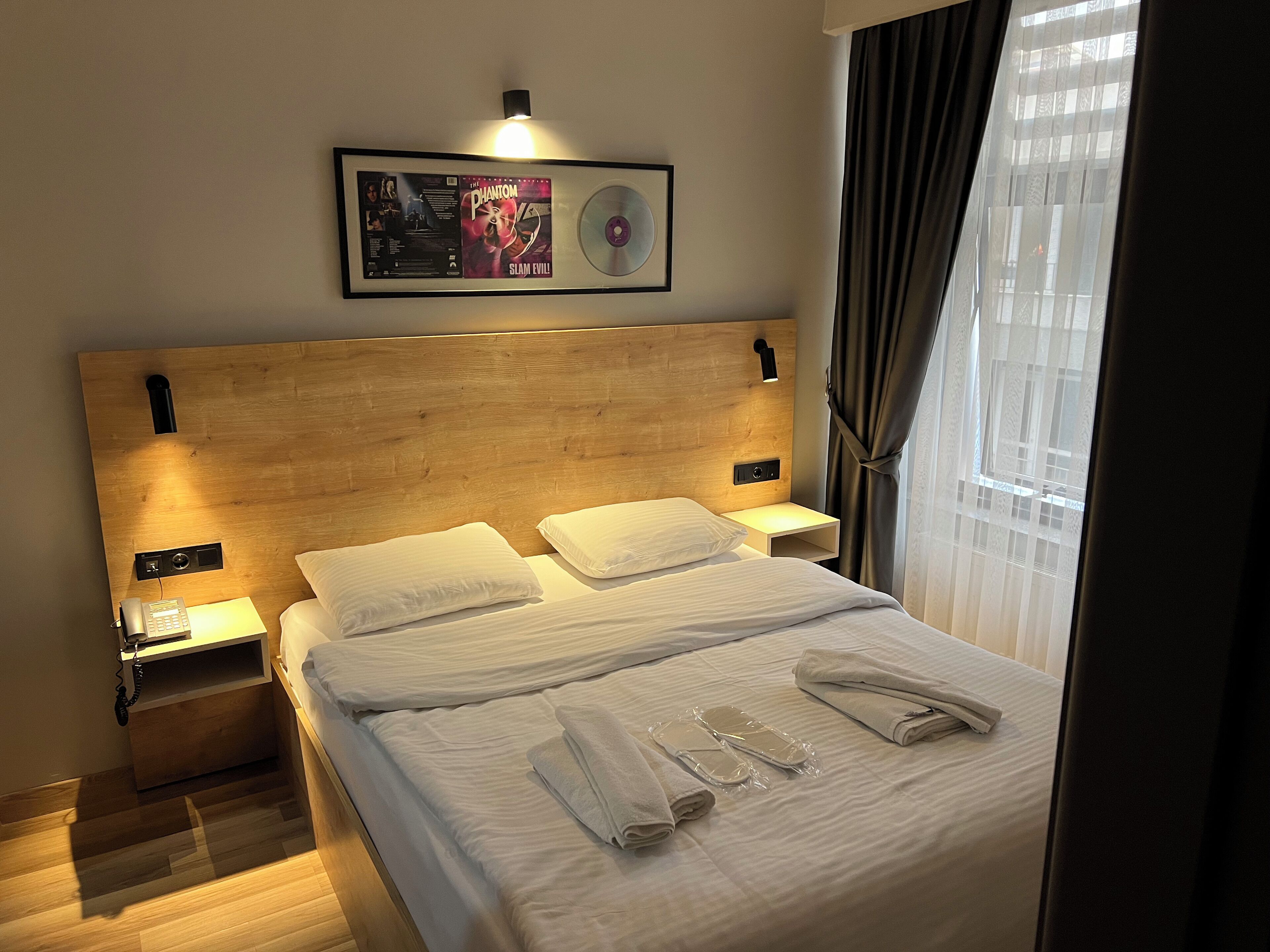 A hotel room with a wooden headboard, two bedside tables with lamps, a framed picture on the wall, and a window with curtains.
