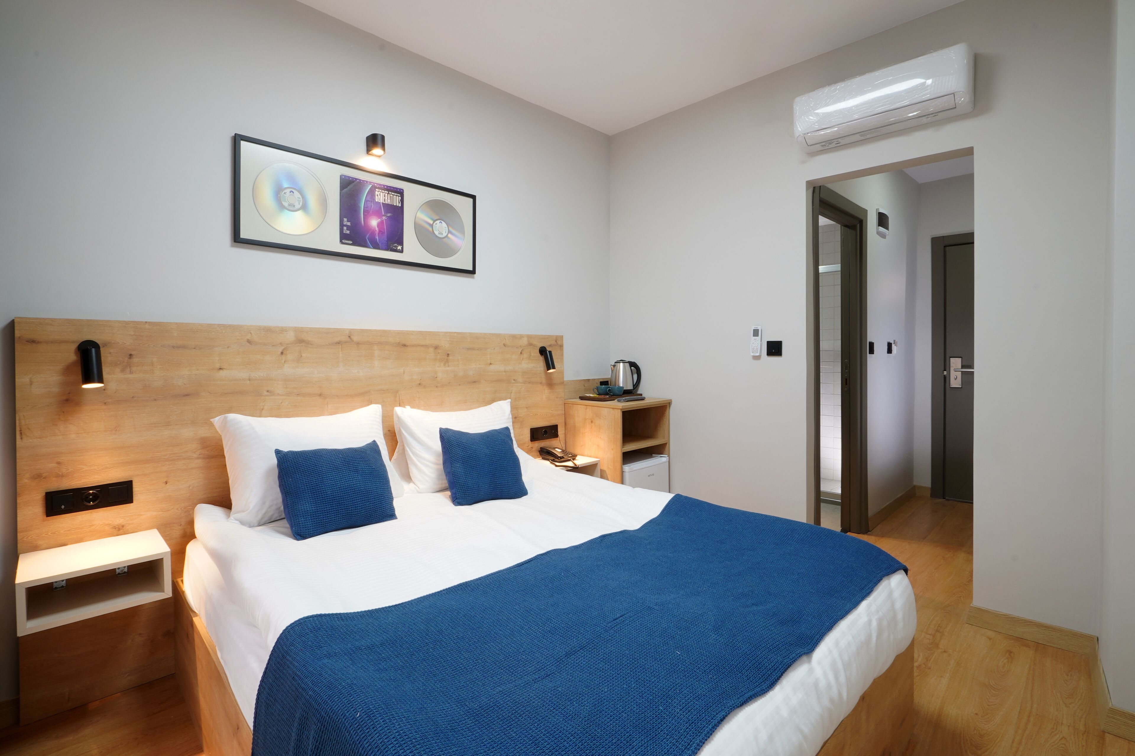 Habitación de hotel ordenada con cabecera de madera, cama con ropa de cama blanca y azul, mesita de noche con lámpara y un cuadro enmarcado en la pared.